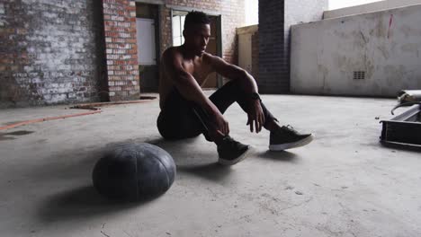 Afroamerikanischer-Mann-Sitzt-Nach-Dem-Training-Mit-Medizinball-In-Einem-Leeren-Städtischen-Gebäude-Und-Ruht-Sich-Aus