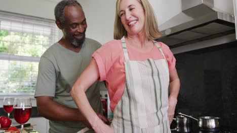 Diverse-senior-couple-putting-on-apron-before-preparing-food-in-kitchen