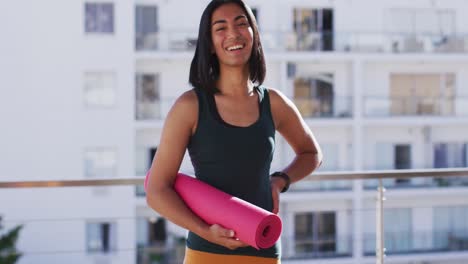 Portrait-of-gender-fluid-male-holding-yoga-mat-smiling-in-the-balcony-at-home