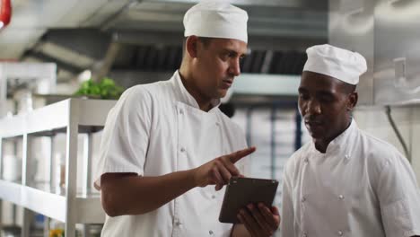 Two-diverse-male-chefs-talking-and-using-tablet-in-restaurant-kitchen