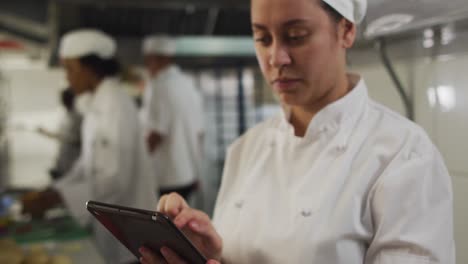 Caucasian-female-chef-using-tablet-in-restaurant-kitchen-with-other-chefs-in-background