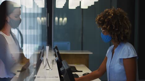 Mixed-race-woman-and-female-receptionist-wearing-face-masks-at-hotel-reception-desk