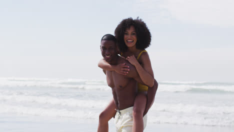 Smiling-african-american-couple-playing-at-the-beach,-man-carrying-woman-piggyback