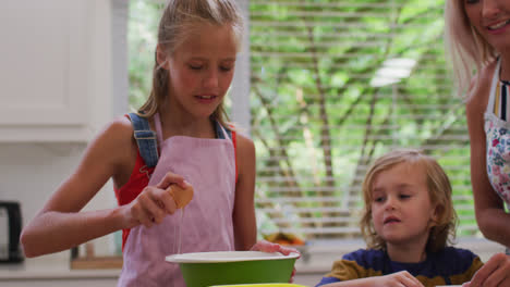 Happy-caucasian-mother-in-kitchen-with-daughter-and-son,-wearing-aprons,-girl-breaking-egg-into-bowl