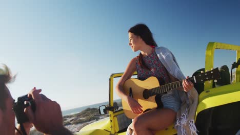 Caucasian-man-taking-pictures-of-woman-sitting-in-beach-buggy-by-the-sea-playing-guitar