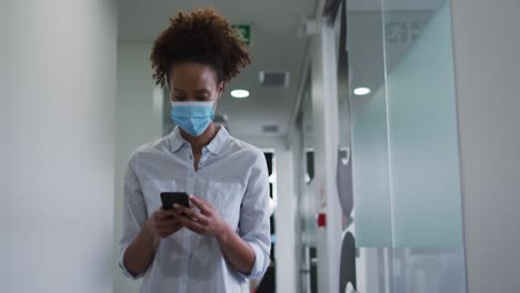 Mixed-race-businesswoman-in-face-mask-using-smartphone-walking-in-corridor-in-office