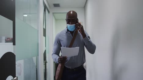 African-american-businessman-in-face-mask-holding-document-talking-on-smartphone-in-office-corridor
