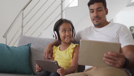Happy-hispanic-father-and-daughter-sitting-on-sofa-using-tablets
