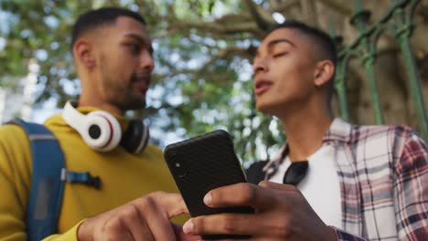 Two-happy-mixed-race-male-friends-standing,-talking-and-using-smartphone-in-the-street