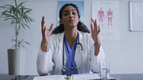 Mixed-race-female-doctor-at-desk-talking-and-gesturing-during-video-call-consultation