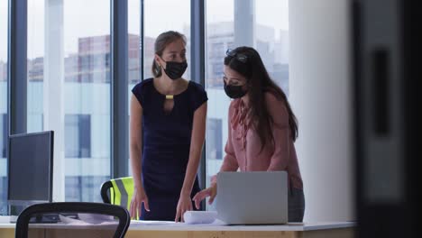 Two-diverse-female-colleague-wearing-face-masks-standing-at-table-looking-at-blueprints-and-talking