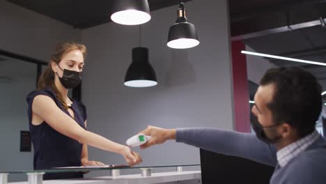 Caucasian-woman-in-face-mask-has-temperature-checked-by-male-colleague-at-office-reception-desk