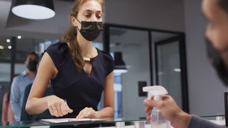 Caucasian-woman-in-face-mask-has-hands-disinfected-by-male-colleague-at-office-reception-desk