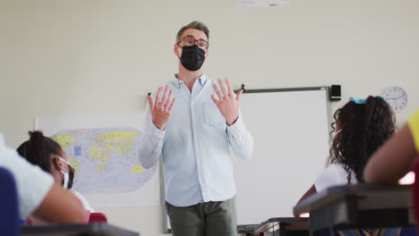Caucasian-male-teacher-wearing-face-mask-teaching-students-in-the-class-at-school