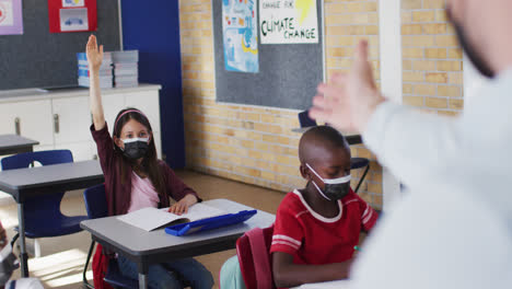 Diverse-schoolchildren-sitting-in-classroom-raising-hands-during-lesson,-all-wearing-face-masks