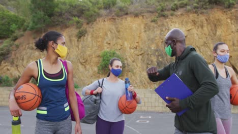 Diverse-female-basketball-team-and-male-coach-wearing-face-masks-greeting-with-elbows