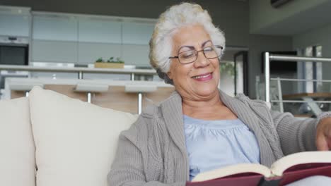 Eine-Afroamerikanische-Seniorin-Liest-Ein-Buch,-Während-Sie-Zu-Hause-Auf-Der-Couch-Sitzt