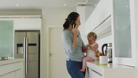 Caucasian-mother-holding-her-baby-talking-on-smartphone-in-the-kitchen-at-home
