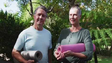 Happy-senior-caucasian-couple-talking-and-smiling-holding-yoga-mats-in-garden