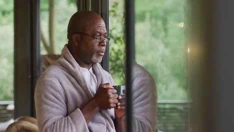 Thoughtful-african-american-senior-man-wearing-bathrobe-drinking-coffee-and-looking-out-of-window