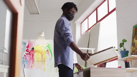 African-american-male-artist-wearing-face-mask-holding-a-painting-and-using-laptop-at-art-studio