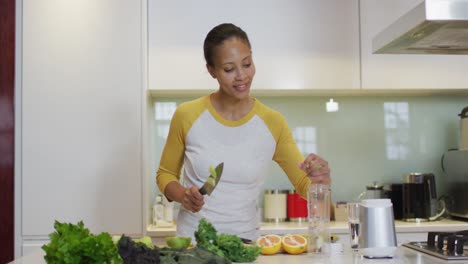Mixed-race-woman-preparing-healthy-drink,-cutting-fruit-and-vegetables-in-kitchen