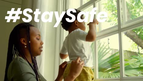 Stay-safe-text-against-african-american-mother-holding-her-baby-near-the-window-at-home