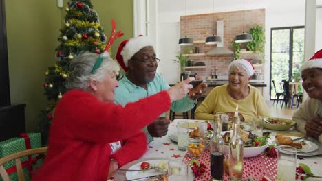 Diverse-senior-friends-in-santa-hats-looking-at-smartphone-and-laughing-at-christmas-dinner-table