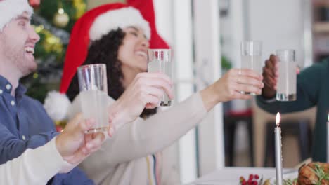 Fröhliche-Gruppe-Verschiedener-Freunde-In-Weihnachtsmützen,-Die-Das-Essen-Feiern-Und-Zur-Weihnachtszeit-Mit-Wein-Anstoßen