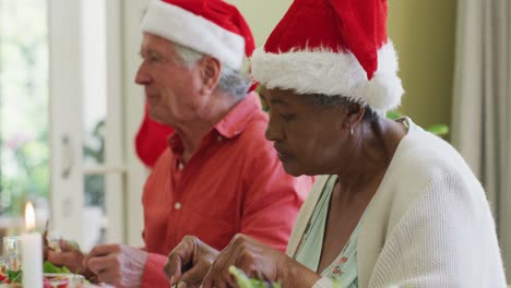 Diverse-senior-man-and-woman-in-santa-hats-listening-and-eating-at-christmas-dinner-table-at-home