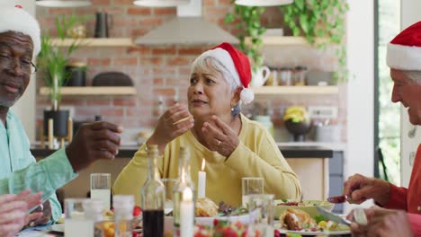 Group-of-happy-diverse-senior-friends-in-santa-hats-talking-at-christmas-dinner-table-at-home