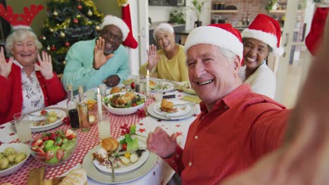 Happy-diverse-senior-friends-in-santa-hats-taking-selfie-and-waving-at-christmas-dinner-table