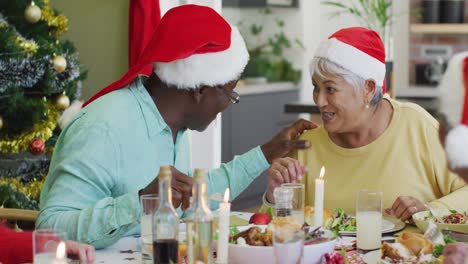Happy-diverse-senior-couple-in-santa-hats-talking-at-christmas-dinner-table-at-home
