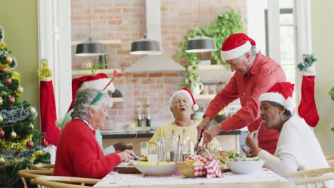 Caucasian-senior-man-in-santa-hat-carving-turkey-at-christmas-dinner-table-with-diverse-friends