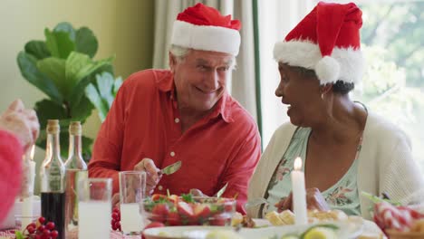 Happy-diverse-senior-couple-in-santa-hats-laughing-at-christmas-dinner-table-at-home