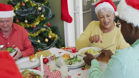 Group-of-happy-diverse-senior-friends-in-santa-hats-passing-food-at-christmas-dinner-table-at-home
