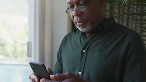 Afroamerikanischer-älterer-Mann,-Der-Zu-Hause-Sein-Smartphone-Am-Fenster-Benutzt