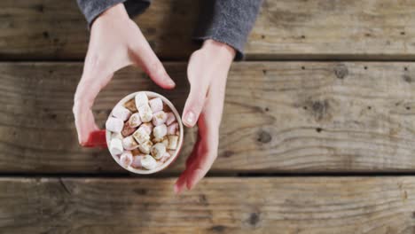 Overhead-view-of-hands-holding-a-hot-chocolate-with-marshmallows-against-wooden-surface