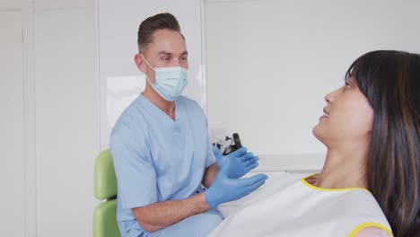 Kaukasischer-Männlicher-Zahnarzt-Mit-Gesichtsmaske,-Der-Eine-Lächelnde-Patientin-In-Einer-Modernen-Zahnklinik-Vorbereitet