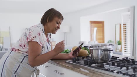 Feliz-Mujer-Mayor-Afroamericana-Preparando-Comida-En-La-Cocina,-Usando-Tableta-Y-Sonriendo