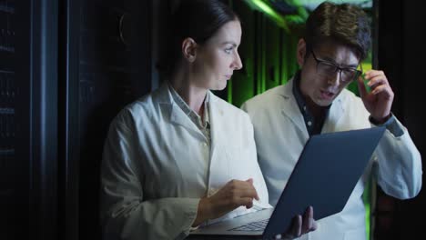 Diverse-female-and-male-it-technicians-in-lab-coats-using-laptop-checking-computer-server