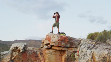 Caucasian-male-survivalist-using-binoculars,-standing-on-mountain-peak-in-wilderness