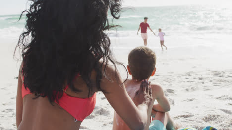 Back-view-of-hispanic-mother-applying-sun-cream-on-son-back-on-beach