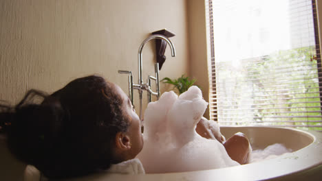 Happy-biracial-woman-with-vitiligo-lying-in-bathtub,-blowing-foam-and-pampering-herself