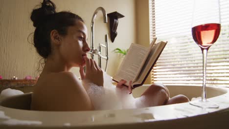Relaxed-biracial-woman-lying-in-bath-with-foam-and-glass-of-wine,-reading-book