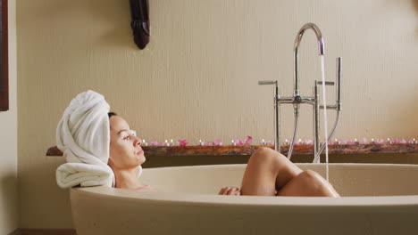 Relaxed-biracial-woman-lying-in-bathtube-and-pampering-herself-with-rose-petals