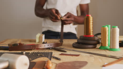Hands-of-african-american-craftsman-using-tool-to-make-a-belt-in-leather-workshop