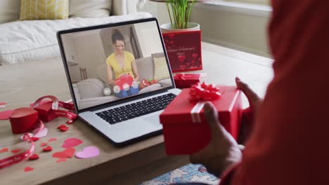 Happy-biracial-woman-opening-gift-and-making-valentine's-day-video-call-on-laptop