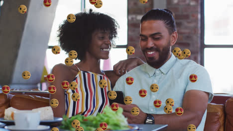 Face-emojis-floating-against-african-american-couple-using-smartphone-sitting-at-a-restaurant
