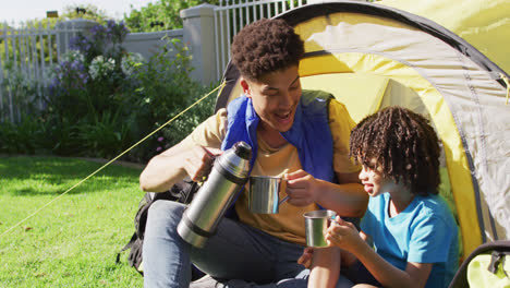 Happy-biracial-man-and-his-son-drinking-from-thermos-flask-in-garden